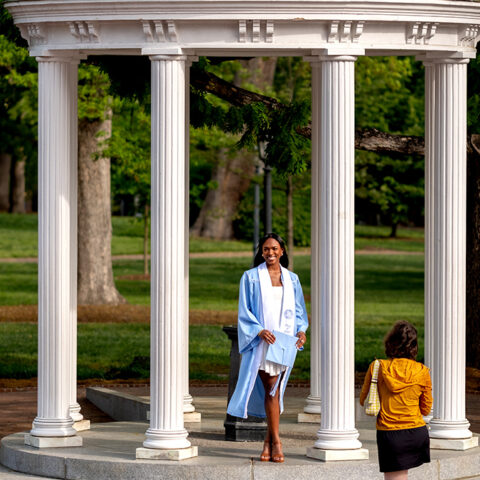 A person in graduate regalia poses for a photo at the Old Well.